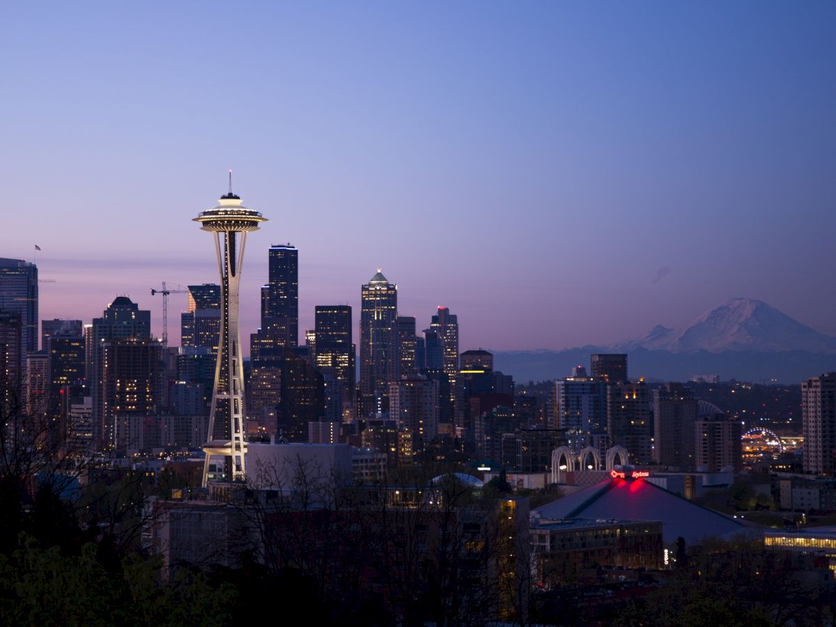 The image shows the Seattle skyline at dusk, featuring the Space Needle prominently, with Mount Rainier visible in the background and city lights illuminating.