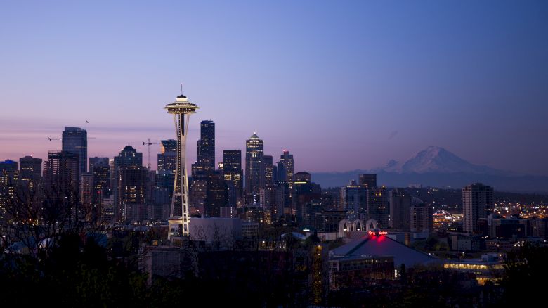 The image shows the Seattle skyline at dusk, featuring the Space Needle prominently, with Mount Rainier visible in the background and city lights illuminating.