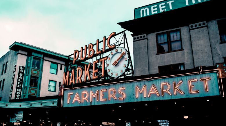 Image shows a neon sign reading "Public Market" with a clock and below it another sign reading "Farmers Market." Buildings frame the sign.