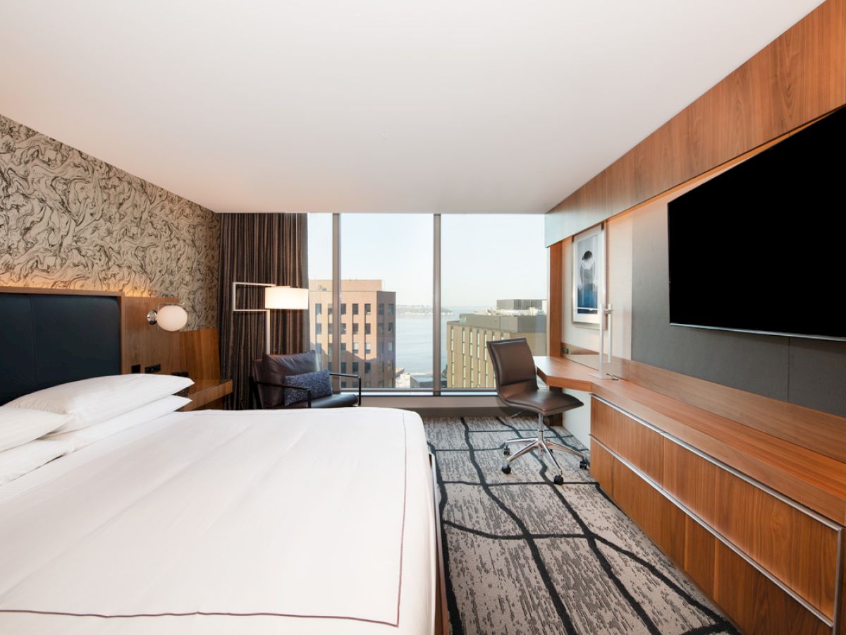A modern hotel room with a large bed, work desk, and a flat-screen TV, featuring city views through floor-to-ceiling windows.