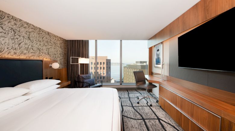A modern hotel room with a large bed, work desk, and a flat-screen TV, featuring city views through floor-to-ceiling windows.