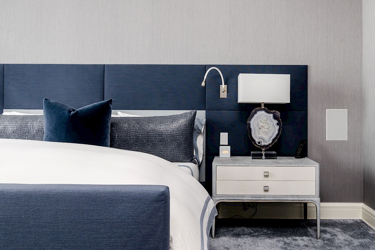 A modern bedroom featuring a blue bed, blue pillows, a white blanket, a white bedside table with a lamp, and a wall-mounted lamp above.