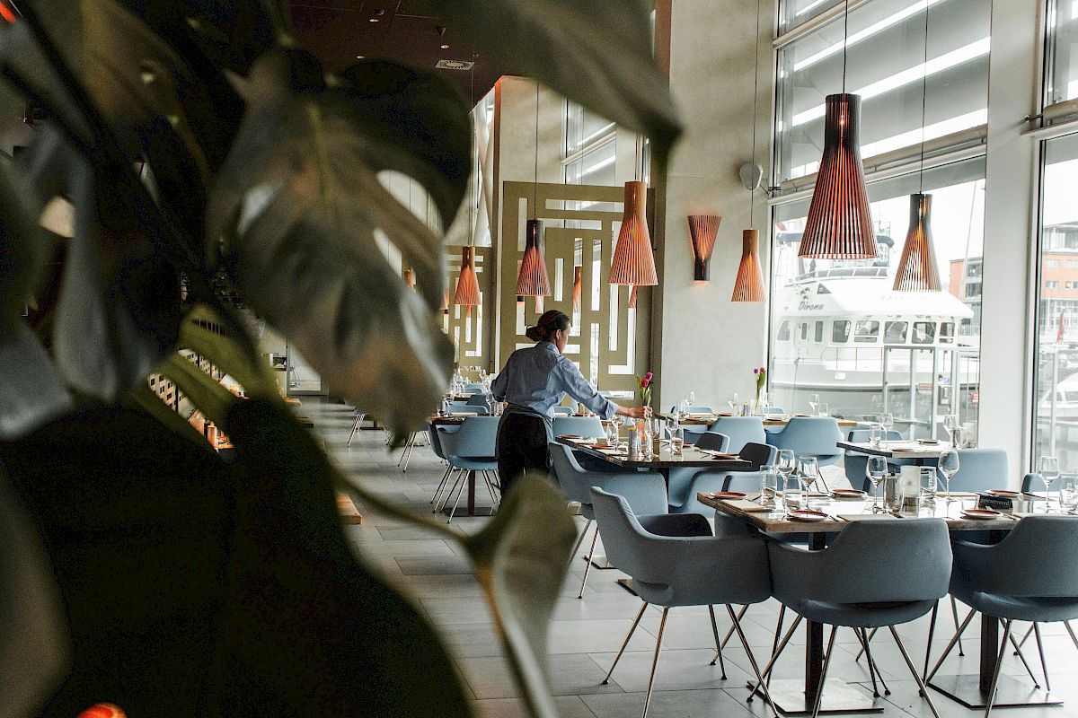 A person is setting up a dining area in a modern restaurant with stylish lighting, blue chairs, and large windows.