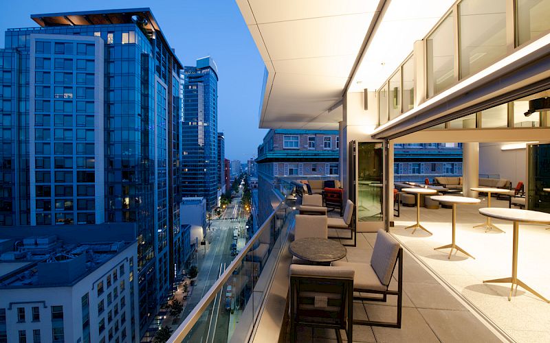 A modern balcony with outdoor tables and chairs overlooks a cityscape with illuminated buildings and streets at dusk.