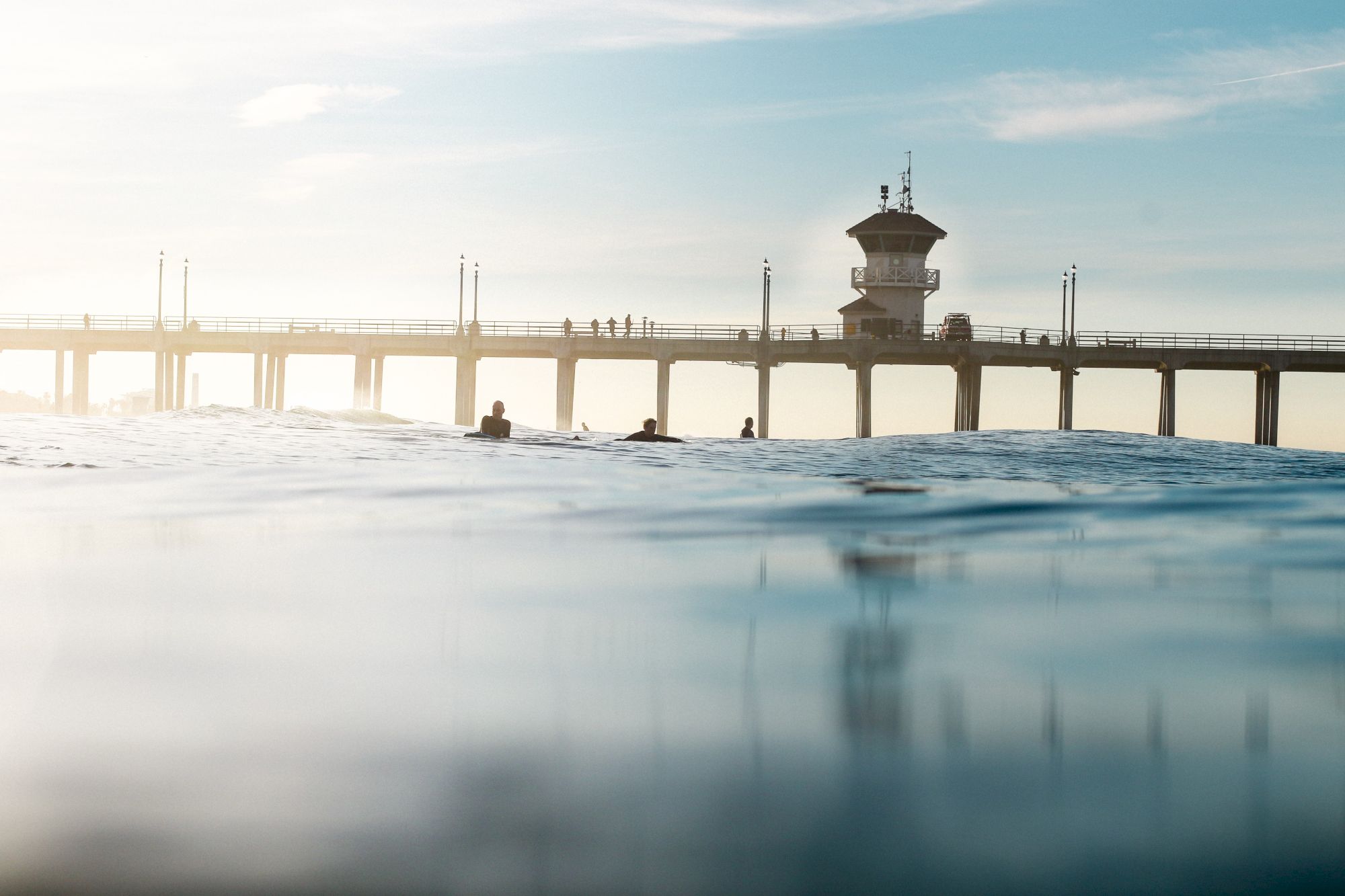 A pier extends over calm ocean waters with a few people walking on it; a tower structure stands on the pier, and some surfers are floating in the water.